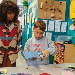 Older brown-skinned woman looks on while a young white boy creates a sculpture